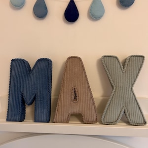 May include: Corduroy letters spelling "MAX" in blue, tan, and green, displayed on a white shelf. Above, a garland of blue teardrop shapes hangs against a white wall, creating a decorative nursery theme.