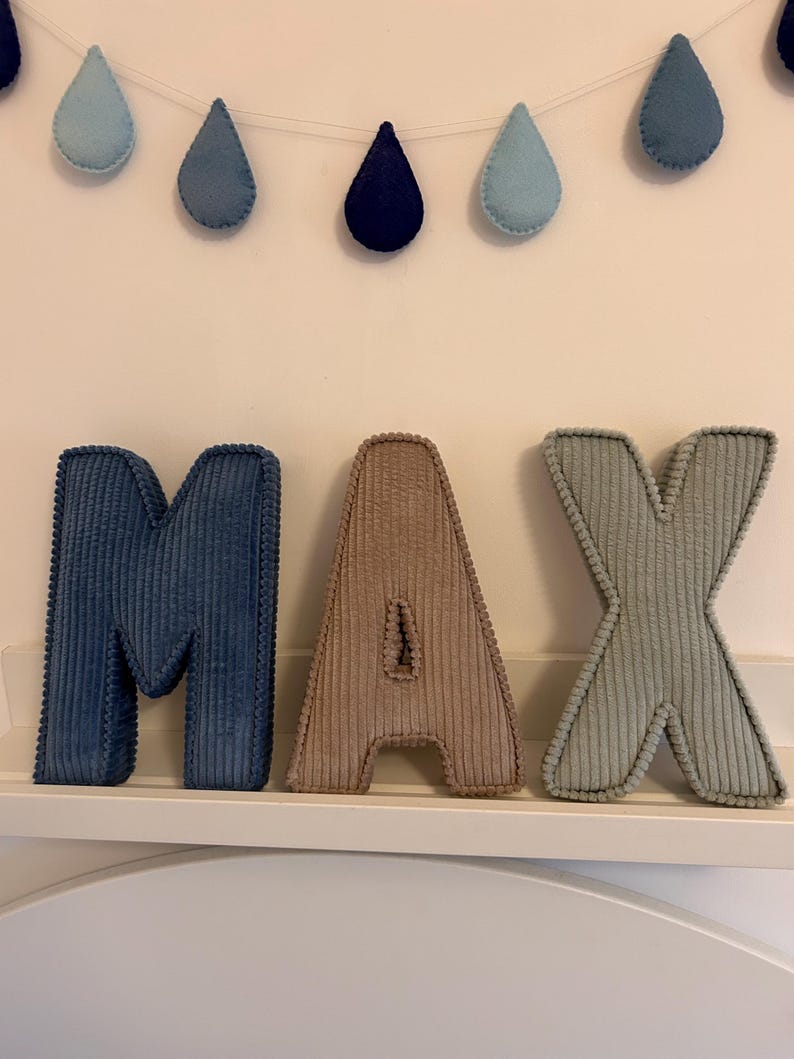 May include: Corduroy letters spelling "MAX" in blue, tan, and green, displayed on a white shelf. Above, a garland of blue teardrop shapes hangs against a white wall, creating a decorative nursery theme.