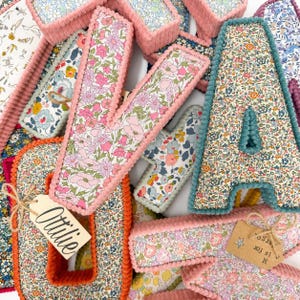 May include: A collection of plush fabric letters featuring various floral patterns in pink, blue, and yellow. The letters have textured corduroy-style edges in matching colors. One letter has a tag reading Ottilie, another has a tag reading M is for Margo.