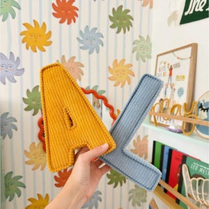 May include: A yellow corduroy letter "A" and a blue corduroy letter "X" are held up in front of a wall with a repeating sun face pattern. The letters are part of a decorative set.