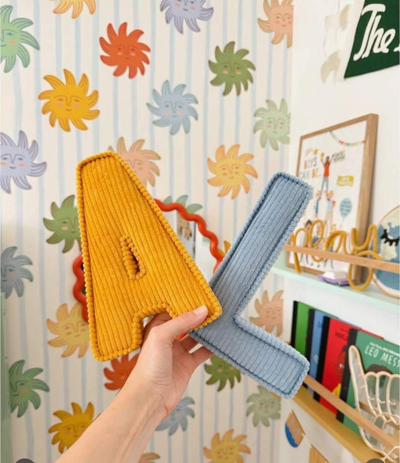 May include: A yellow corduroy letter "A" and a blue corduroy letter "X" are held up in front of a wall with a repeating sun face pattern. The letters are part of a decorative set.