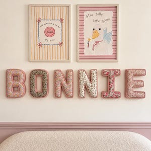 Op de afbeelding: Zes met stof beklede muurletters die het woord BONNIE vormen in diverse bloemenpatronen met roze biezen. Daarboven hangen twee ingelijste prints: een met gele strepen en een hartje met de tekst How sweet it is to be loved by you, en een met roze strepen en een gans met de tekst stay silly little goose.