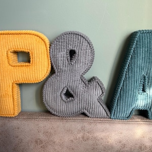 May include: Three corduroy letters: a yellow "P", a gray ampersand, and a teal "A". The letters are plush and have a ribbed texture. They are arranged on a textured surface against a light green wall.