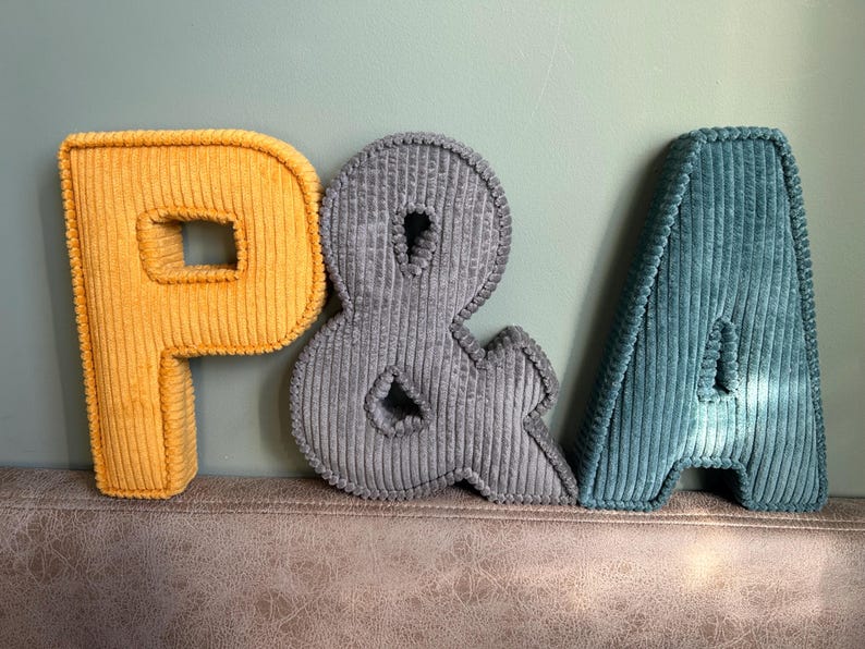 May include: Three corduroy letters: a yellow "P", a gray ampersand, and a teal "A". The letters are plush and have a ribbed texture. They are arranged on a textured surface against a light green wall.