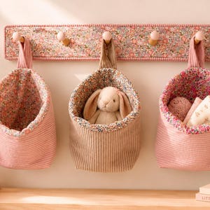 Puede incluir: Un perchero de pared de madera con cinco ganchos, cubierto con una tela floral rosa. Tres bolsas de almacenamiento de tela colgantes están suspendidas de los ganchos. Las bolsas tienen exteriores de pana rosa y forros florales, con un conejo de peluche.