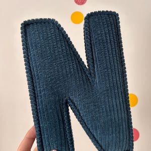 May include: A teal corduroy letter "N" with a textured surface and a stitched border. The letter is displayed against a white background with colorful circular decorations.