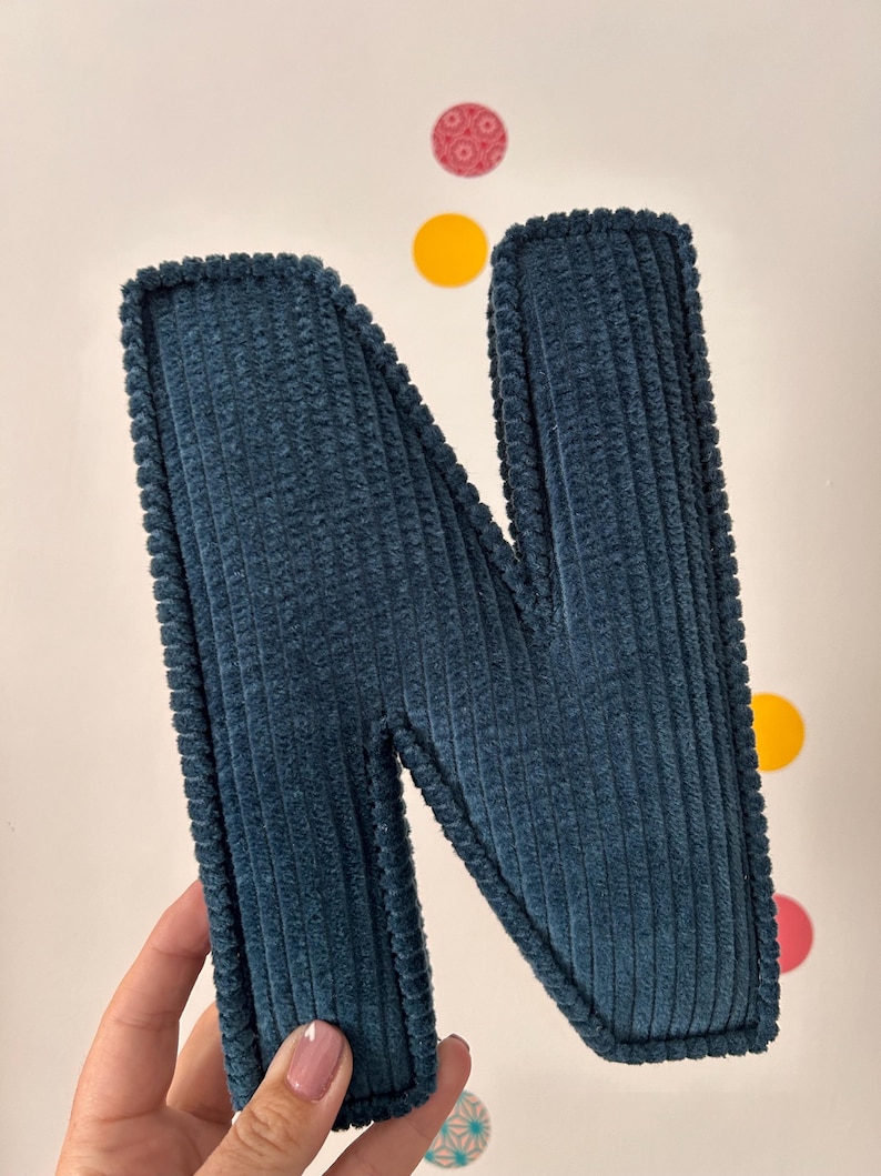 May include: A teal corduroy letter "N" with a textured surface and a stitched border. The letter is displayed against a white background with colorful circular decorations.