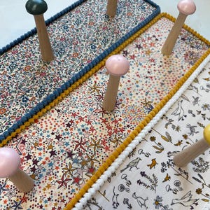 Puede incluir: Tres percheros de pared rectangulares con paneles cubiertos de tela con patrones distintos: un estampado floral, un motivo de estrellas y un dise&ntilde;o de naturaleza con p&aacute;jaros y setas. Cada perchero tiene ganchos de madera con puntas de color.