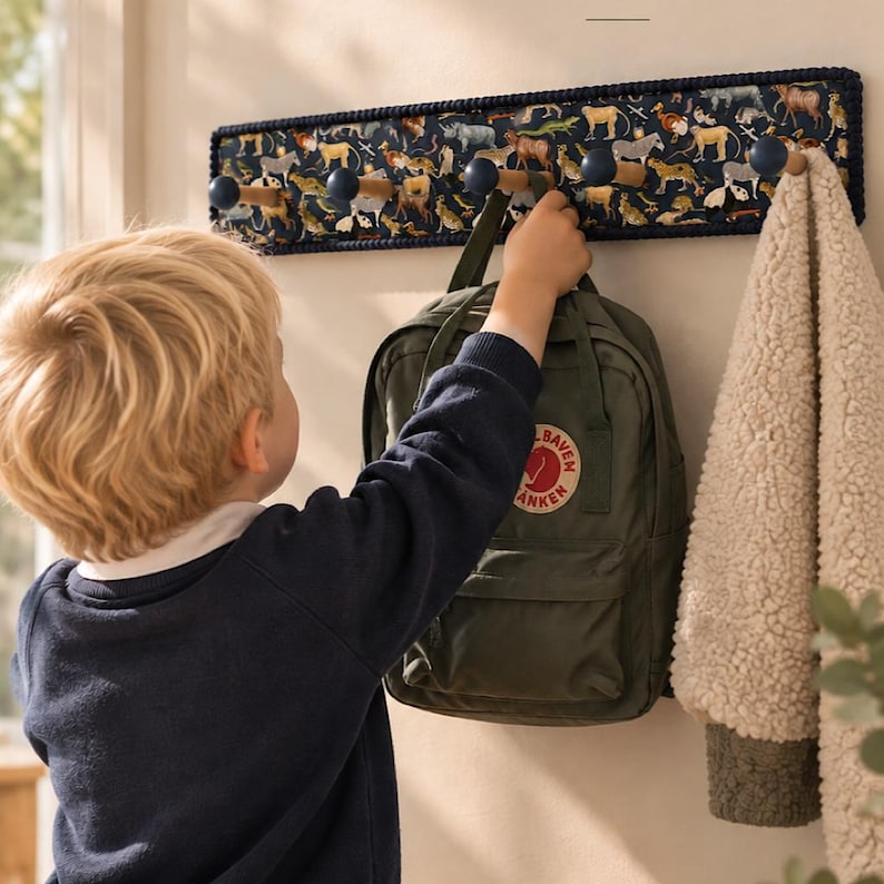 Puede incluir: Un perchero de pared rectangular con un panel de tela azul oscuro y un estampado de animales multicolor. El perchero incluye cinco ganchos de madera con extremos redondeados en azul oscuro, que sostienen una mochila verde y una chaqueta de forro polar beige.