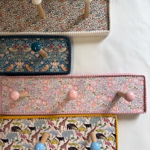 May include: A collection of decorative wall-mounted coat racks. Each rack features a rectangular wooden base covered in patterned fabric, with wooden pegs for hanging items. The fabrics include floral and animal prints, with colorful trim and knobs.