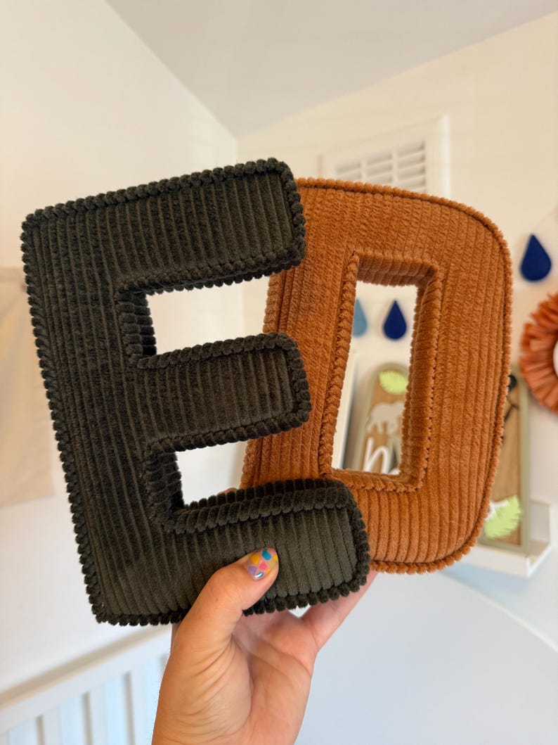 May include: Two corduroy letters, "E" in dark green and "D" in orange, are displayed. The letters are plush and have a ribbed texture. The background includes a white wall and decorative elements.