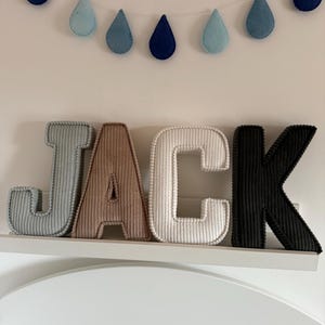 May include: A decorative wall display featuring the name "JACK" in corduroy letters. The letters are in shades of gray, brown, white, and black. Above the letters is a garland of felt raindrops in blue hues.