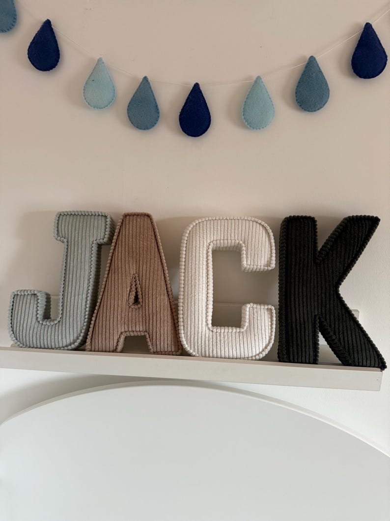 May include: A decorative wall display featuring the name "JACK" in corduroy letters. The letters are in shades of gray, brown, white, and black. Above the letters is a garland of felt raindrops in blue hues.