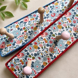 Puede incluir: Dos percheros rectangulares de pared con un estampado floral de flores azul claro, rosas y amarillas sobre fondo blanco. Cada perchero tiene tres ganchos de madera, algunos pintados de rosa claro. Los bordes est&aacute;n rematados con hilo azul o rojo.