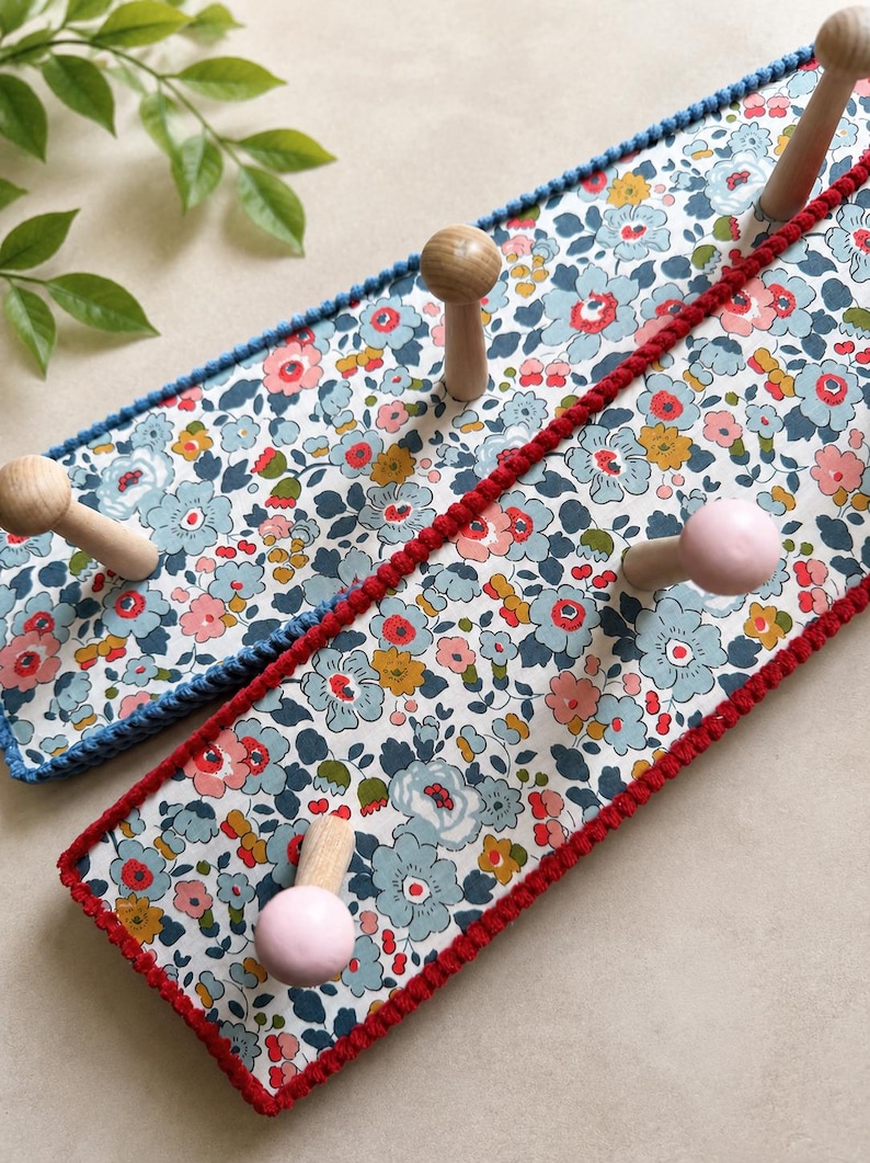 Puede incluir: Dos percheros rectangulares de pared con un estampado floral de flores azul claro, rosas y amarillas sobre fondo blanco. Cada perchero tiene tres ganchos de madera, algunos pintados de rosa claro. Los bordes est&aacute;n rematados con hilo azul o rojo.