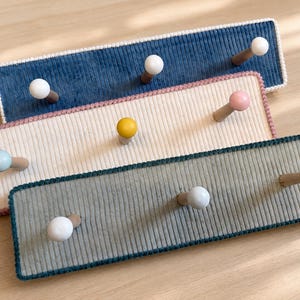 May include: Three rectangular wall-mounted coat racks featuring ribbed fabric surfaces in blue, cream, and grey. Each rack has three wooden pegs with rounded, coloured tips in white, yellow, pink, and light blue, finished with a crocheted border.