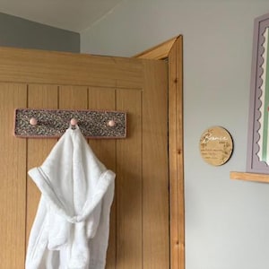 Puede incluir: Un perchero de pared rectangular con un borde de ganchillo rosa y un centro de tela moteada multicolor. El perchero cuenta con tres peque&ntilde;os ganchos de madera redondos de color rosa claro. Una bata blanca con capucha cuelga del gancho central.