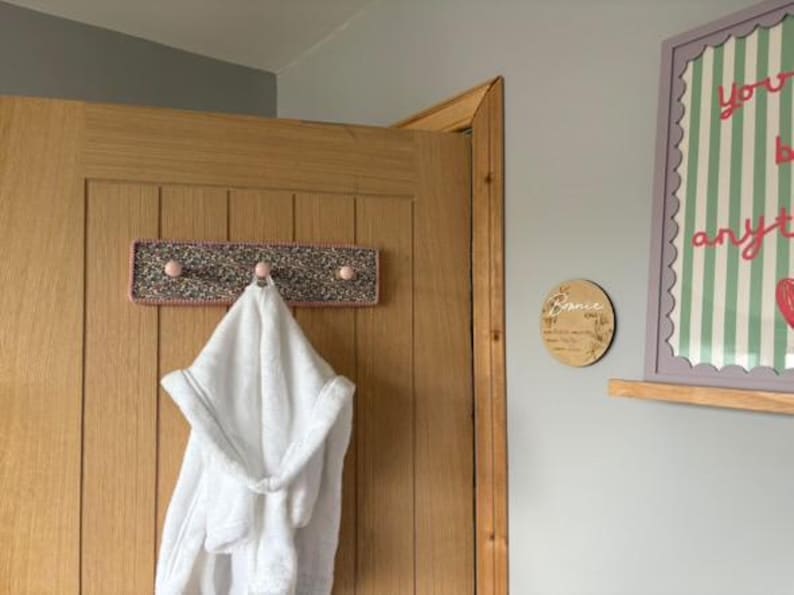 Puede incluir: Un perchero de pared rectangular con un borde de ganchillo rosa y un centro de tela moteada multicolor. El perchero cuenta con tres peque&ntilde;os ganchos de madera redondos de color rosa claro. Una bata blanca con capucha cuelga del gancho central.