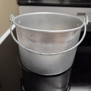 May include: A cylindrical aluminum bucket with a smooth, matte silver finish. The bucket features a rounded rim and a sturdy, hinged wire bail handle attached to two small metal loops on opposite sides of the upper exterior wall.