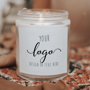 Pu&ograve; includere: Una candela bianca in un barattolo di vetro trasparente con un'etichetta bianca che dice "YOUR logo DESIGN OR TEXT HERE". La candela &egrave; appoggiata su un tappeto marrone e bianco con un motivo.