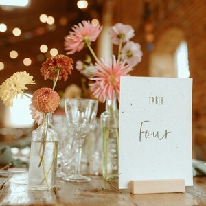 Op de afbeelding: Een witte kaart met de tekst "TABLE FOUR" erop, gesteund op een houten standaard. De kaart is omringd door een boeket roze, gele en oranje bloemen in glazen vazen.