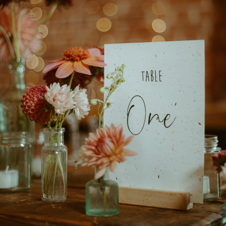 Op de afbeelding: Een witte tafelnummerkaart met zwarte tekst die "TABLE One" luidt, wordt getoond op een houten standaard. De kaart is omringd door kleine glazen vazen gevuld met roze en witte bloemen.