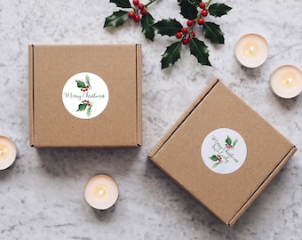 Può includere: Due scatole quadrate in cartone marrone con adesivi circolari bianchi decorati con illustrazioni di agrifoglio ad acquerello. Un adesivo riporta la scritta Merry Christmas, l'altro Merry Christmas love Emily. Le scatole sono circondate da agrifoglio e candele.