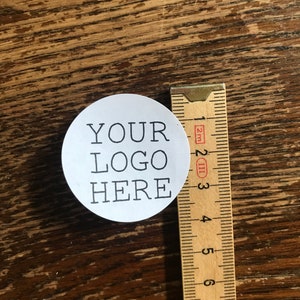 May include: A white round sticker with black text that says "YOUR LOGO HERE" next to a wooden ruler.