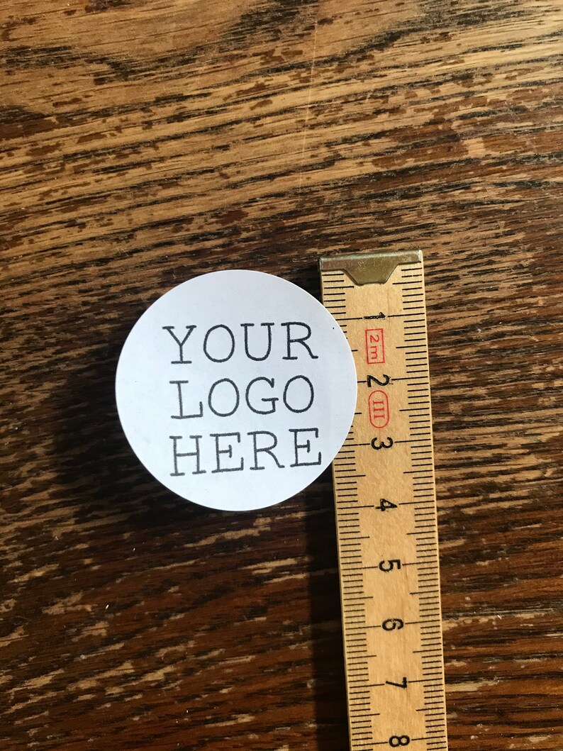 May include: A white round sticker with black text that says "YOUR LOGO HERE" next to a wooden ruler.