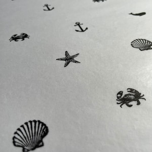 May include: A white surface is covered with black illustrations of nautical elements. The illustrations include anchors, starfish, seashells, crabs, and whales. The design is repeated across the surface, creating a pattern.