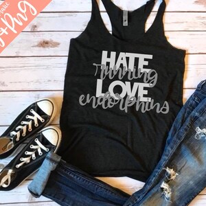 May include: A black tank top with the text "Hate Running Love Endorphins" in white lettering. The tank top is laying on a white wooden surface with a pair of black sneakers and blue jeans.