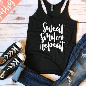 Może przedstawiać: Czarny top na ramiączkach z napisem "Sweat Smile + Repeat" w białych literach. Top leży na białym drewnianym tle z parą czarnych butów sportowych i niebieskimi dżinsami.