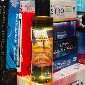 May include: A clear glass bottle of Belladonna's Botanicals Spiritual & Emotional Healing Bath & Body Oil. The label lists ingredients and instructions. The bottle is 4 fl oz. The background includes books and oracle cards.