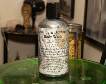 May include: A silver body wash bottle with a black cap, labeled "Crossing & Hexbreaking Body Wash" by Belladonna's Botanicals. The label lists ingredients and the net weight of 8 fl oz. The bottle sits on a green glass surface.