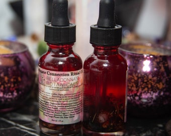 Könnte beinhalten: Zwei kleine Glasflaschen gefüllt mit einer roten Flüssigkeit und Kräutern. Die Flaschen haben schwarze Tropfer und Etiketten mit der Aufschrift "Ultimate Connection Ritual Oil" und "BellaDonna's Botanicals".