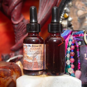 May include: Two glass bottles of ritual oil with black droppers. The bottles are filled with a dark red liquid and have labels that read "Come to Me Ritual Oil" and "Belladonna's Botanicals". The bottles are on a white surface.