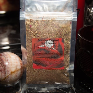 May include: A small, clear plastic bag filled with dried herbs and flowers. The bag has a red label that reads "Marriage Proposal Spell Powder BellaDonna's Botanicals". The label also includes a list of ingredients and instructions for use. The bag is 0.8 oz.