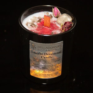 Op de afbeelding: Een zwarte kaars pot met een rode was top en een label met de tekst "Lucifer Devotional Candle". De kaars is versierd met gedroogde rozenblaadjes, kristallen en een houten lont.