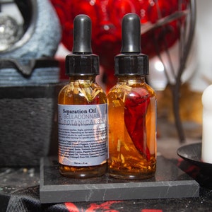 Può includere: Due bottiglie di vetro di Separation Oil di Belladonna's Botanicals, ciascuna con un contagocce nero. Una bottiglia contiene un peperoncino rosso. Le etichette includono testo. Le bottiglie sono su una superficie di marmo nero.