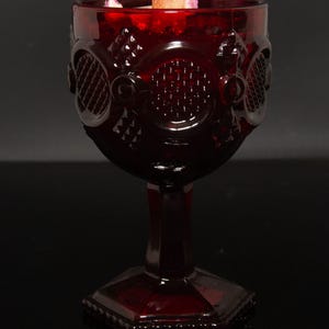 May include: A deep red glass goblet candle holder featuring a textured, embossed geometric pattern on the exterior bowl. The vessel stands on a hexagonal base and contains a candle with a visible wooden wick and small decorative floral elements.
