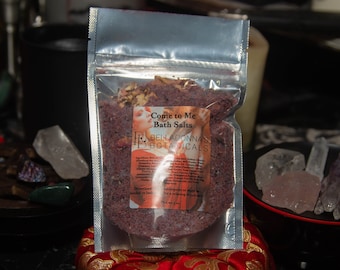 Puede incluir: Una pequeña bolsa resellable llena de sales de baño moradas. La etiqueta dice "Come to Me Bath Salts" y "Bella Donna's Botanicals". La bolsa está sobre una superficie roja.