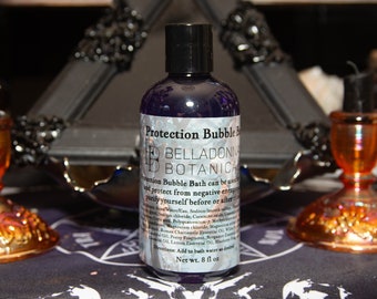 May include: A purple bottle of Protection Bubble Bath by Belladonna Botanica. The bottle is labeled with the product name and instructions for use. The bottle is sitting on a black surface with a gold and orange decorative object in the background.