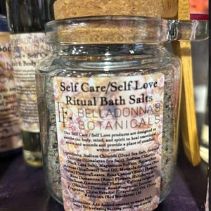 Pode incluir: Um frasco de vidro transparente com tampa de cortiça cheio de sais de banho rituais. O rótulo diz "Self Care/Self Love Ritual Bath Salts" e lista os ingredientes. O frasco contém 142 gramas.