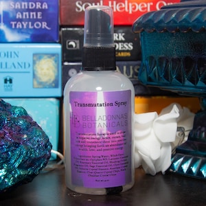 Może przedstawiać: Przezroczysta butelka ze szkła z czarną końcówką rozpylacza i białą etykietą. Etykieta zawiera napis "Transmutation Spray" i "BellaDonna's Botanicals". Butelka jest wypełniona przezroczystym płynem i stoi na drewnianej powierzchni.