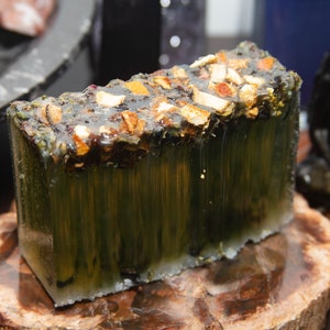 Peut inclure: Une barre de savon verte et brune avec des morceaux de fruits et d'herbes incorporés. Le savon est posé sur une surface en bois.