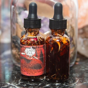 May include: Two small amber glass bottles with black dropper tops filled with a dark liquid and dried herbs. The label on the bottle reads "Marriage Proposal Spell Ritual Oil" and "BellaDonna's Botanicals".