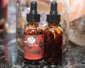 Könnte beinhalten: Zwei kleine bernsteinfarbene Glasflaschen mit schwarzen Tropfern, gefüllt mit einer dunklen Flüssigkeit und getrockneten Kräutern. Das Etikett auf der Flasche lautet "Marriage Proposal Spell Ritual Oil" und "BellaDonna's Botanicals".
