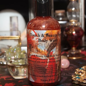 May include: A clear bottle with a black lid filled with a glittery orange liquid. The label reads "Youth & Beauty Spell Body Wash" and "BELLADONNA BOTANICA". The bottle is 8 fl oz.