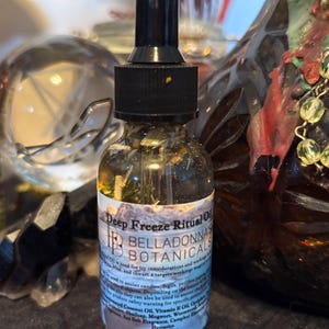 May include: A clear glass dropper bottle containing a golden liquid with visible botanical herbs inside. The white label features the text Deep Freeze Ritual Oil and Belladonna Botanicals, with a black dropper cap and a black base.