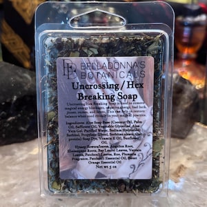 May include: A rectangular bar of Uncrossing/Hex Breaking Soap by Belladonna's Botanicals in clear plastic packaging. The soap is blue and topped with dried herbs and botanicals. The label lists ingredients and states a net weight of 5 oz.